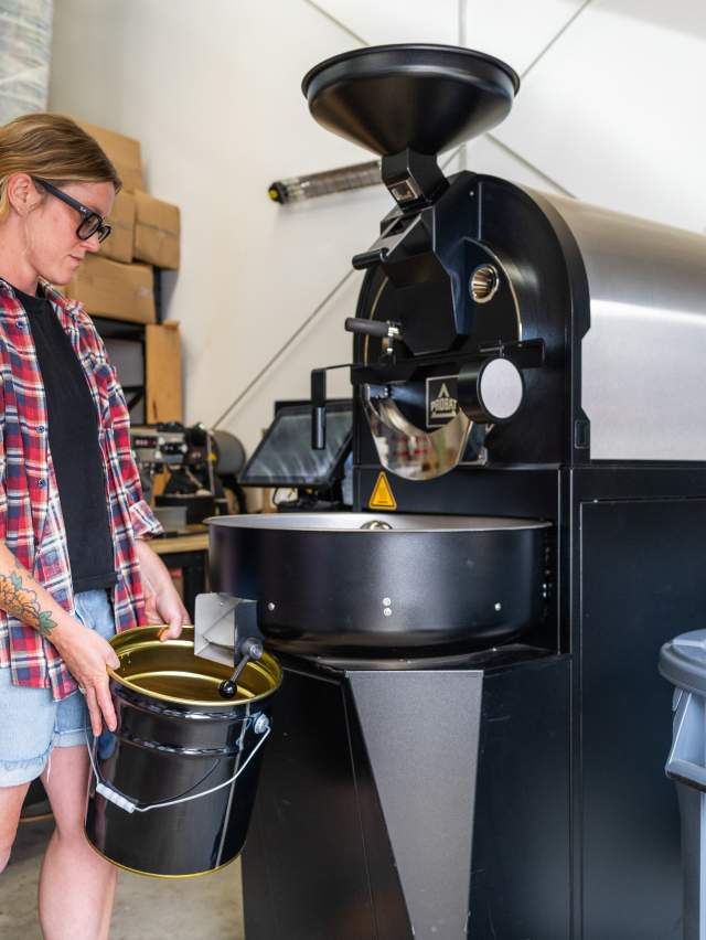 A woman in a casual clothes stands next to a coffee roaster catching beans in a tin