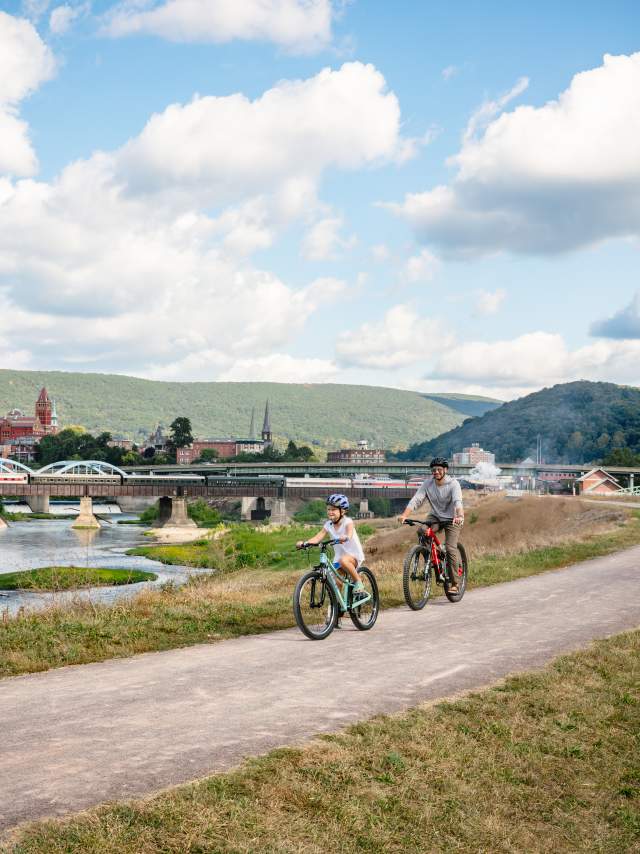 Adult and child riding bicycles along the C&O Canal Towpath in Cumberland, Maryland, with a river, bridge, downtown buildings, and forested mountains in the background under a blue sky.