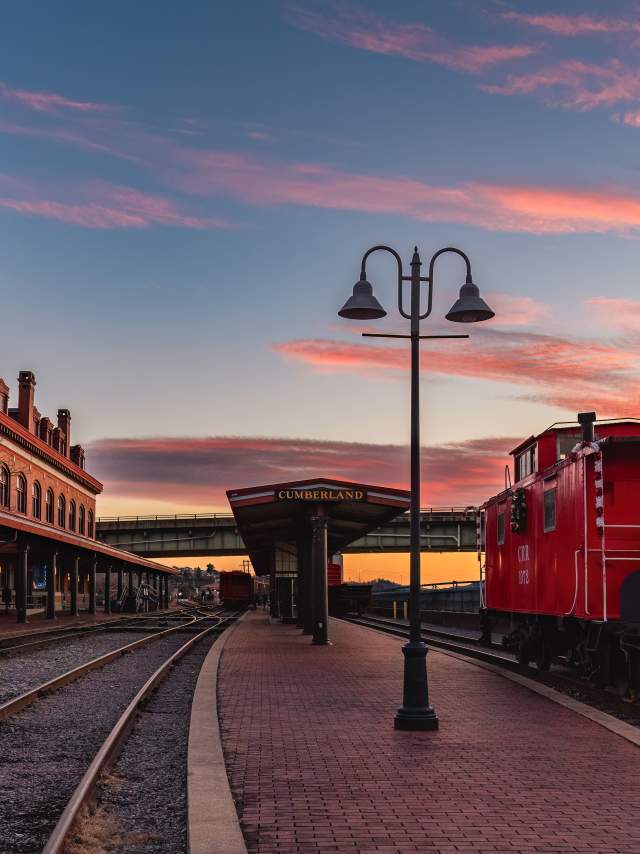 A red train caboose with a holiday wreath sits parked on train tracks in front of a historic train station at sunset.