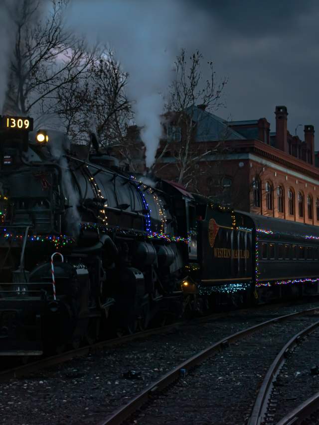 A steam engine decorated with Christmas lights and a holiday wreath on the engine is parked in front of a historic brick depot at night.