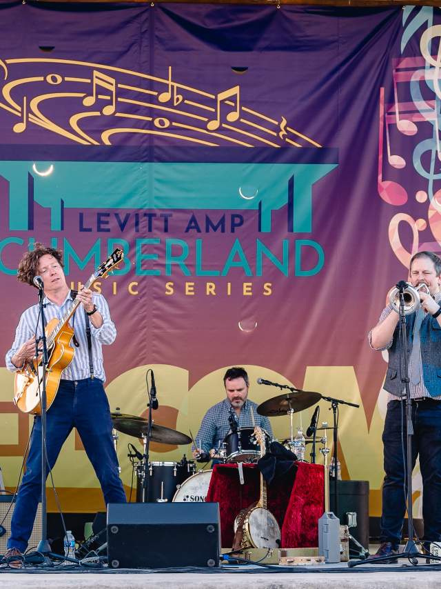 Full band on stage playing music in front of a colorful backdrop that says "Levitt AMP Cumberland Music Series."