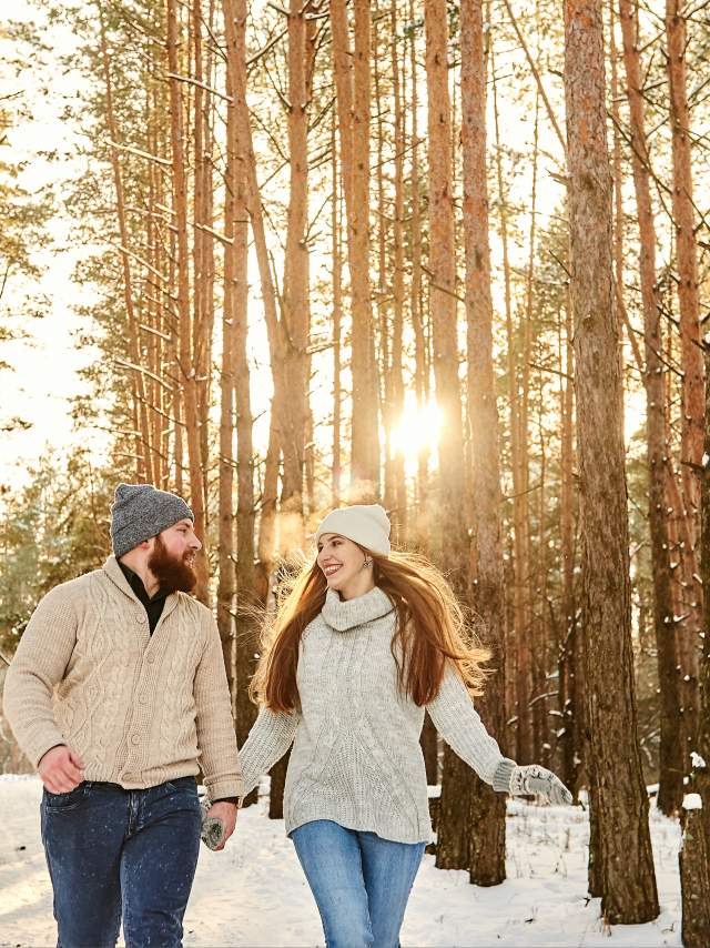 A man and woman hold hands and walk through the snow covered forest.