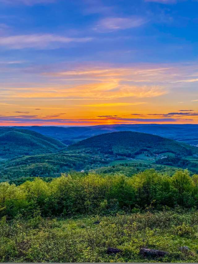 The sky is painted in orange, yellow, purple and blues as the sun sets over the mountains along the grassy mountain edge.