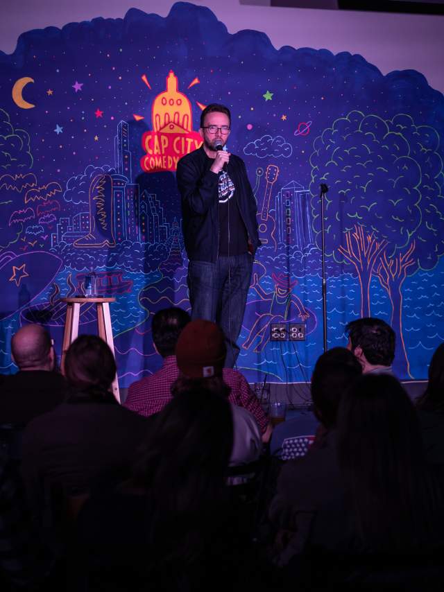 Joe List performing stand-up comedy on the stage of Cap City Comedy Club in front of a shadowy audience.