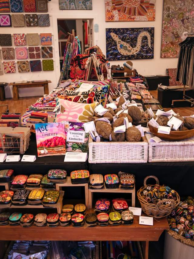 Art and crafts display at Artlandish Aboriginal Art Gallery in Kununurra - colourful artworks, small goods such as cushion covers and carved boab nuts are visible.