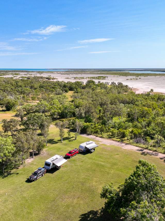 Aerial view of cars and caravans in the grassed campground at Banana Well Getaway, with the ocean visible in the background