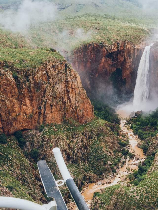 The foreground shows the step rail of the helicopter where this image is taken and one side of the helicopter windows. In the far distance Revolver Falls waterfall spills water over a rock face into a muddy rushing creek below. There is mist coming off the waterfall and lush vegetation all around