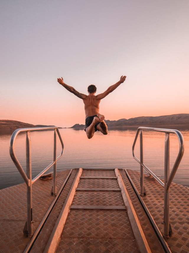 A person is shown jumping from a boat into Lake Argyle at sunset; his arms are raised, and his legs tucked in.