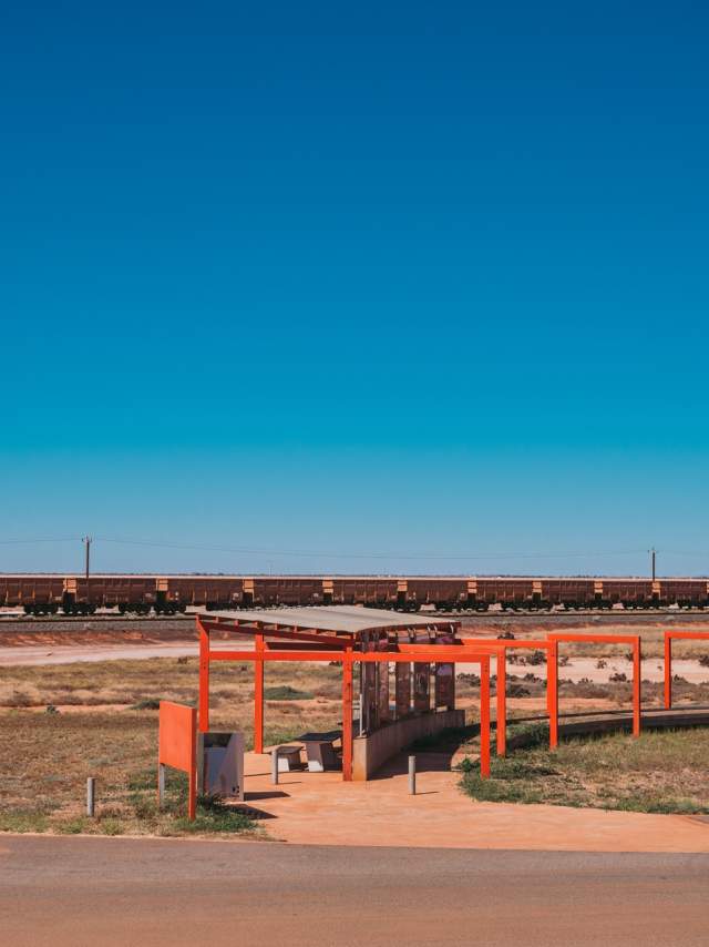A car is parked at Redbank Bridge Lookout with a long iron ore train visible in the background. Alongside the carpark is a shaded lookout area with intrepretative signage.