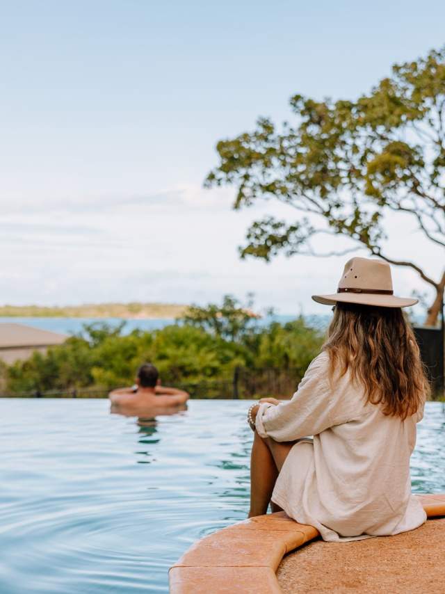 Looking from the swimming pool across to the waters of Cygnet Bay, a man is inside the pool, with a woman sitting on the edge. Both are looking at the view.