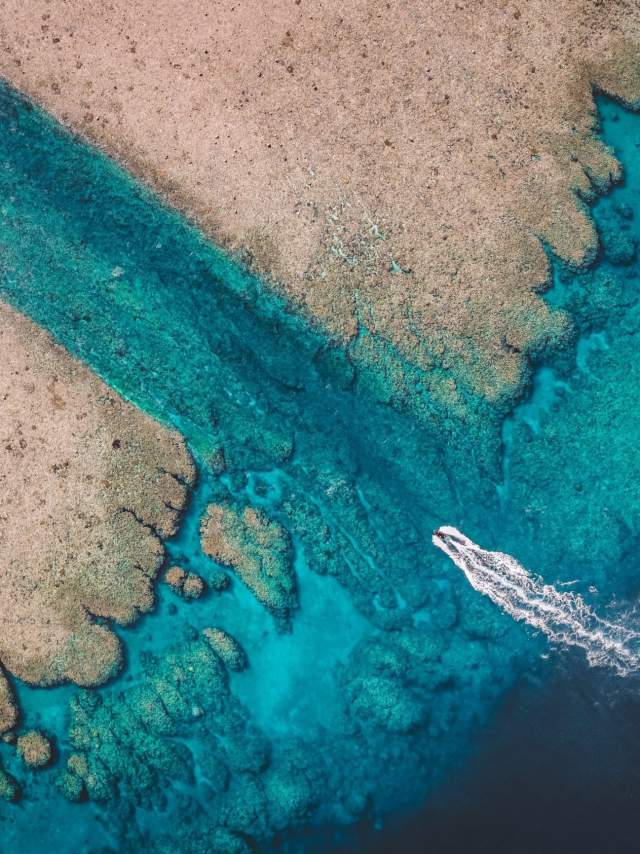 Shot from overhead, a tender vessel is seen entering a narrow channel at the Rowley Shoals. You can see trails of water from the vessels engine, deep blue water outside the channel and lighter blue in the channel.