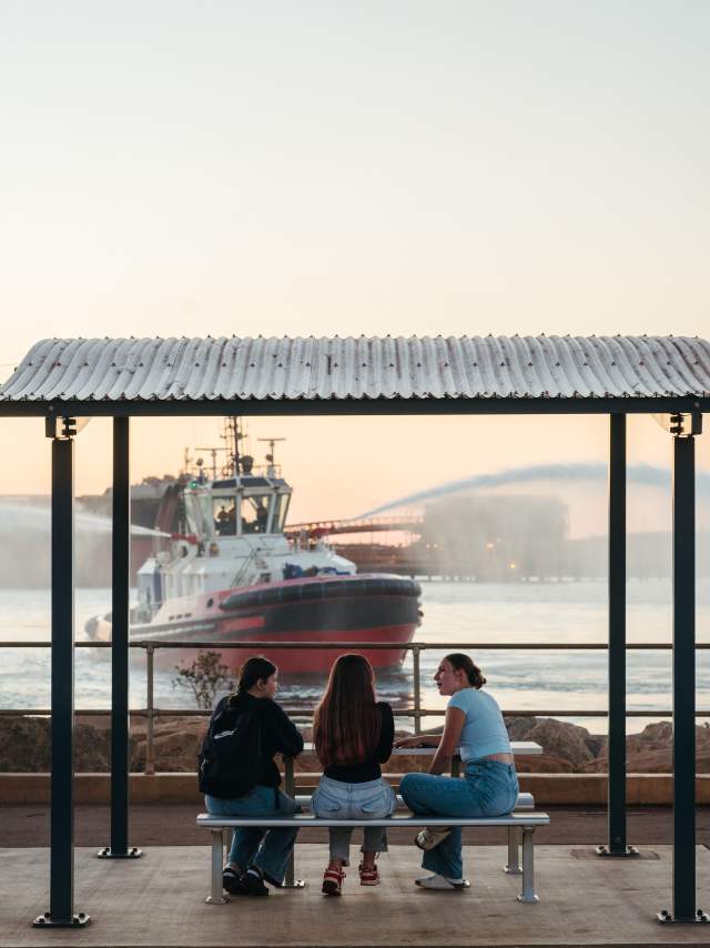 Three people sitting at a picnic table at Marapikurrinya Park with a harbour tug vessel working in the background.