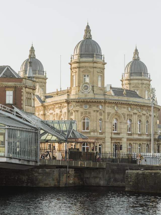 Hull's Maritime Museum in the background. The water of Princes Quay in the foreground. The sky is hazy at sunrise.