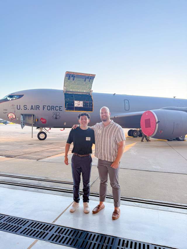 Two people stand side by side on an airport tarmac in front of a large U.S. Air Force aircraft. One has their arm around the other, and both are smiling at the camera. The plane behind them has an open hatch and visible “U.S. AIR FORCE” lettering on its side. The sky is clear and bright, giving the photo a clean, sunny backdrop