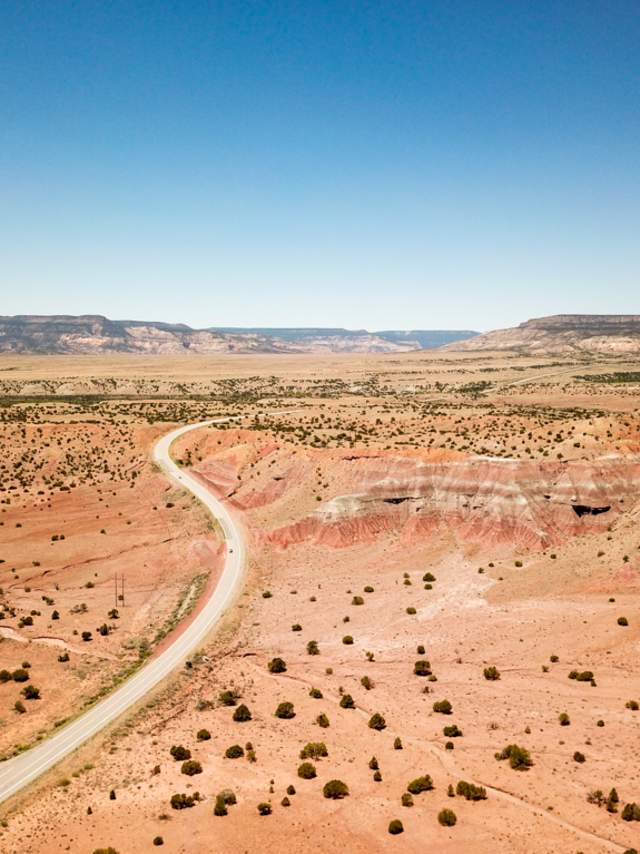 a two lane road stretches across a vast desert landscape with shubbery, plateaus rise out of the landscape in the distance