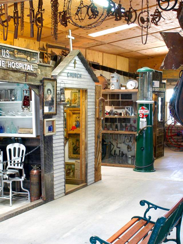 A rustic room filled with vintage maritime and industrial artifacts is seen. A structure labeled "U.S. Marine Hospital" with various objects and a chair occupies one side. Nearby, a small chapel-like display labeled "Church" is present. Old tools and machinery parts hang from the ceiling and walls.