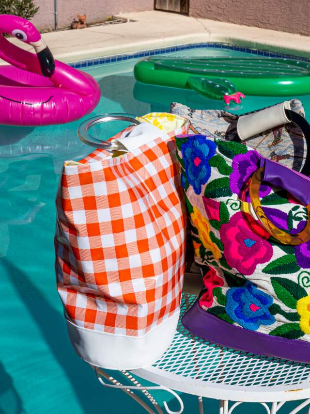 A colorful poolside scene featuring a vibrant, floral-patterned bag and a checked towel on a white metal table, with inflatable pool rings floating in the background.