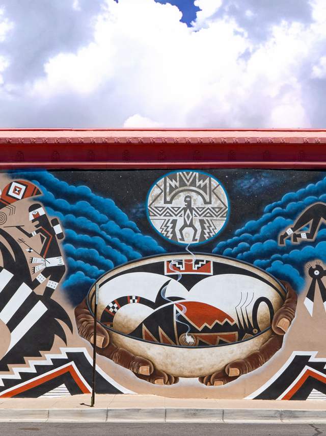 A vibrant mural featuring intricate Native American motifs and patterns. A person stands in front of the mural, emphasizing its grand scale.
