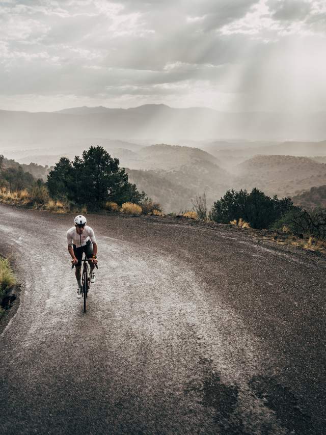 A person is cycling on a winding road through a mountainous landscape with trees and rocky terrain surrounding them. Sunbeams break through the cloudy sky, casting light on the scene.