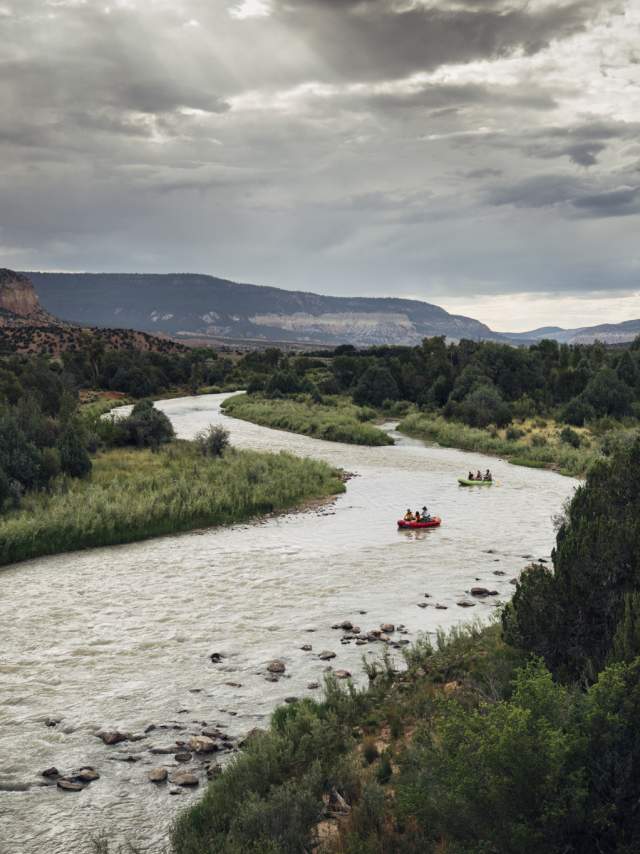 Rafters navigate a winding river through a scenic desert landscape under a cloudy sky.