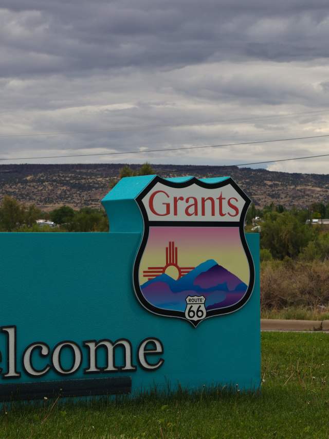 Welcome sign for Grants, featuring the Route 66 emblem, set against a backdrop of a cloudy sky and distant hills.