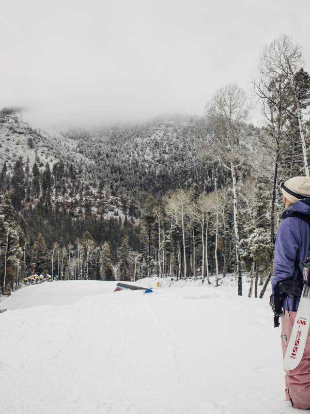 A person wearing winter clothing stands on a snowy slope, holding skis branded with "Line." They are looking towards a snow-dusted mountain landscape with tall trees around. Some snow-covered structures and ski equipment are visible in the distance.