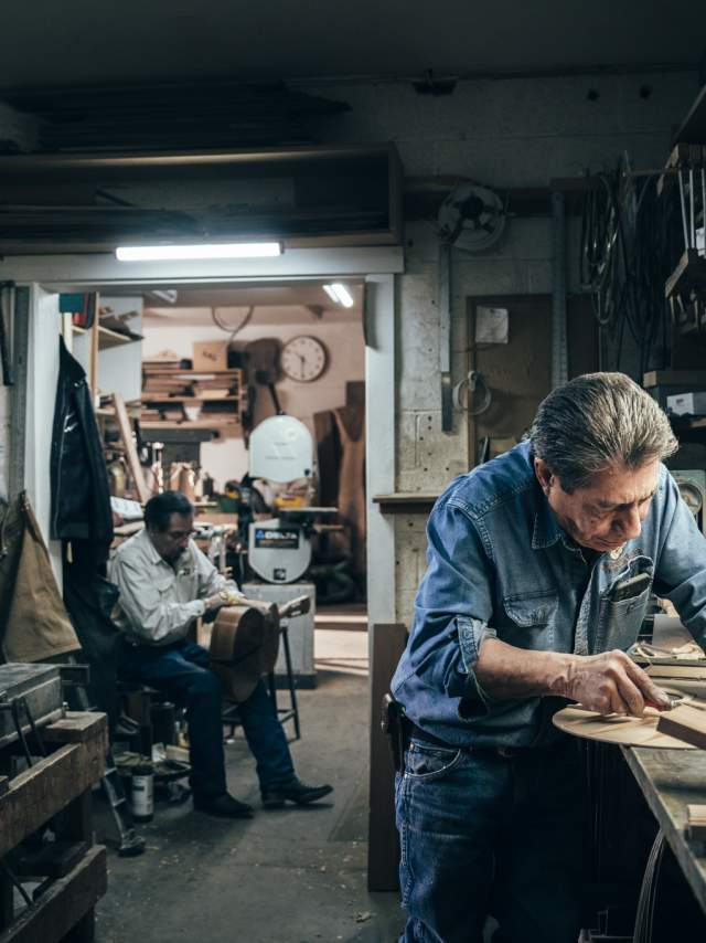 A person is working intently on a piece of wood in a cluttered workshop, while another person sits in the background holding the body of a guitar.