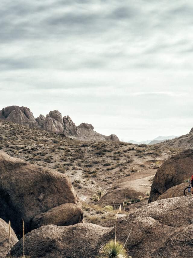 Two people with bicycles standing on a rocky terrain in a desert landscape, with mountains in the background under a cloudy sky.