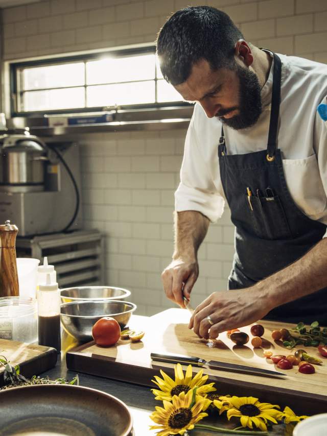 A chef diligently prepares food in a professional kitchen. The countertop is adorned with various fresh ingredients, including herbs and vegetables.