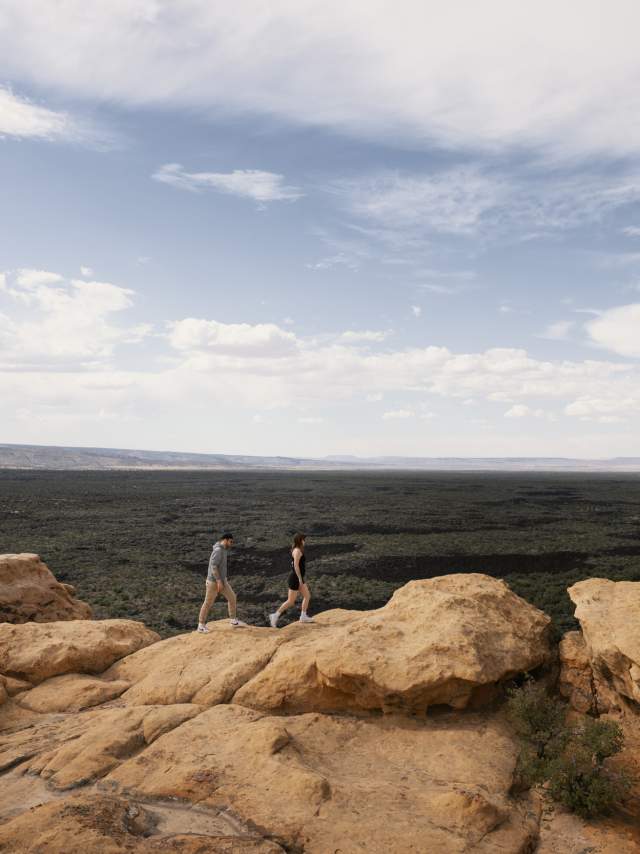 Two people hiking on rocky terrain in a vast desert landscape under a cloudy sky.