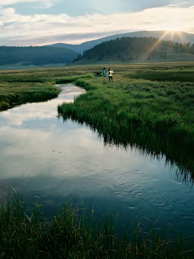 Two people stand by a small creek which flows between a grass meadow, featuring rolling hillsides and the sun shining in the background