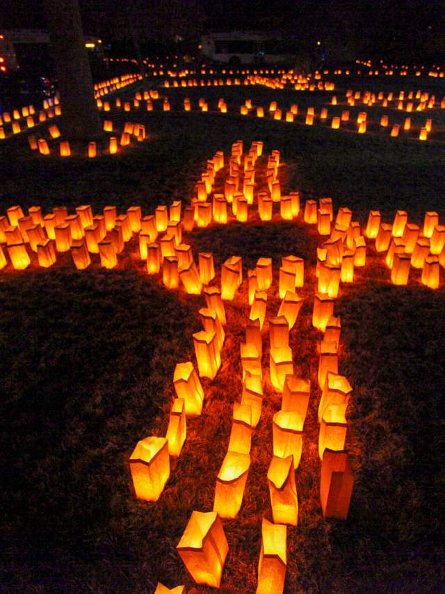 Nighttime view of numerous luminarias arranged to form a large cross, illuminating the area with a soft, warm glow.