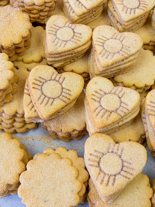 Piles of biscochito cookies, some with a traditional rounded shape and some in a heart shape branded with a Zia symbol.