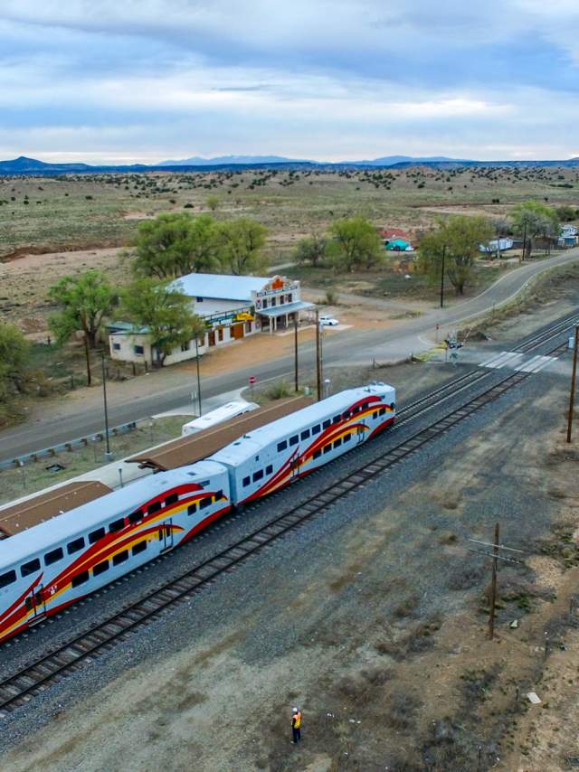 a line of train cars with a red roadrunner painted on the side wait at a station along a solitary stretch of tracks running across a green plain