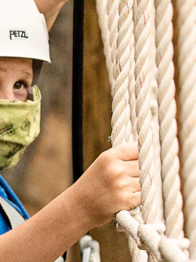 A boy safely climbs a ropes course with his mask on.