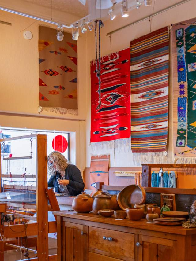 a woman stands behind a wooden loom, a wooden dresser is in the fore ground and there are weaved rugs adorning the walls