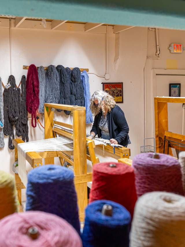 colorful spools of thread are in the foreground as a woman weaves on a wooden machine, there are loose bundles of yarn hanging from the wall behind her