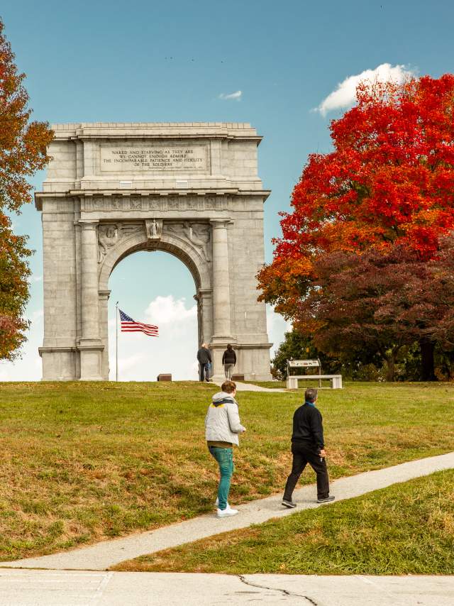 An older male and female walk casually up a hill in front of the monument arch at the Valley Forge National Historic Park surrounded by trees with fall foliage.