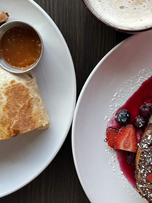 An egg chorizo burrito sits on a plate next to house potatoes while another plate nearby features mascarpone-stuffed French toast covered in fresh berries and berry compote at Greenhouse Cafe in Kalamazoo.