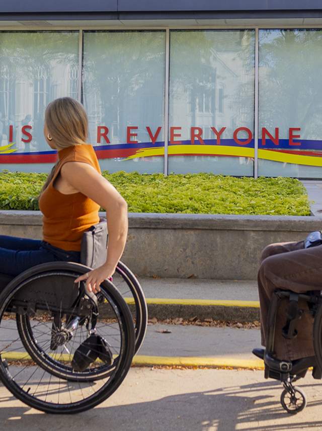 Two young women in wheelchairs roll in front of the Kalamazoo Institute of Arts with a painting on the window that says "Art is for Everyone."