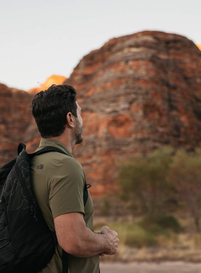 A man stands on a walking track, looking up at the striped domes of the Bungle Bungle Range, Purnululu National Park