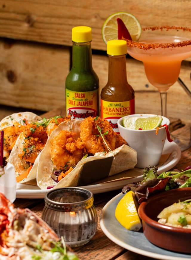 A view of a fully laden table with crab, tacos and other plates of seafood, with a cocktail glass, at Crabshack seafront restaurant on the Sussex coast.