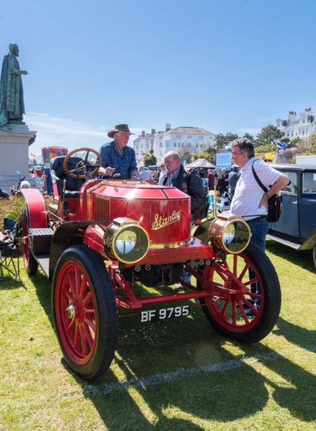 A group of men stand round a red vintage car parked on the green next to a statue in the seaside town of Eastbourne in Sussex