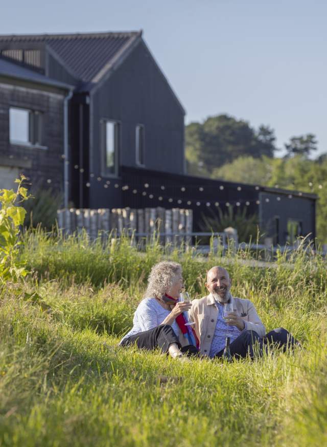 A couple sit in the vines with a glass of wine at Tillingham vineyard, with black barn buildings behind