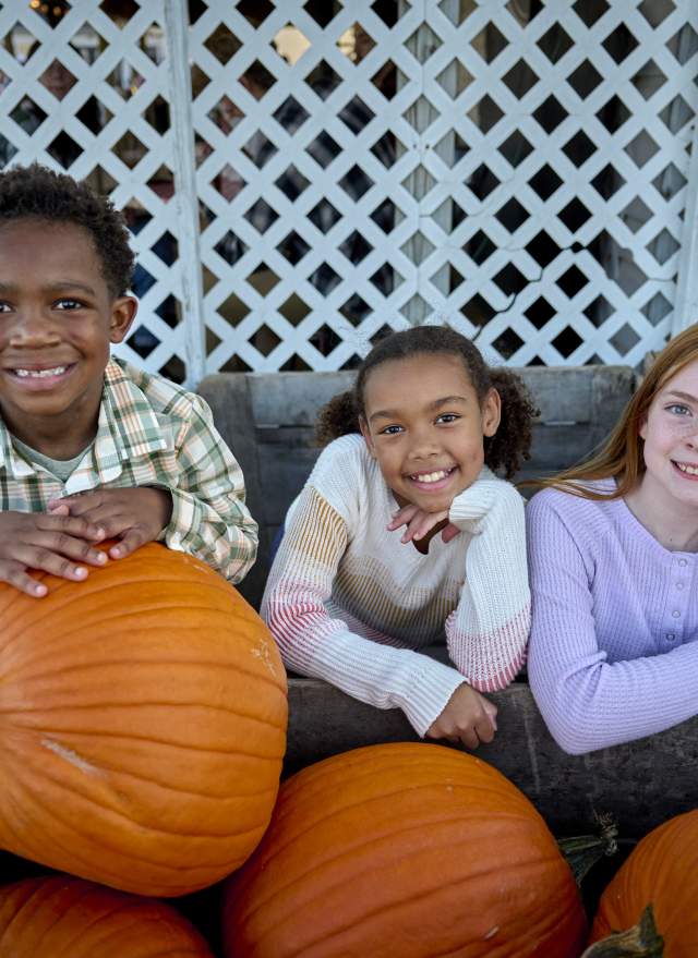 Three kids leaning on pumpkins, smiling