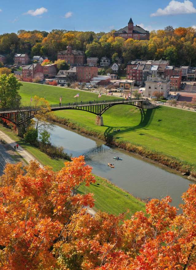 A picturesque view of Galena, Illinois, in fall, featuring a scenic bridge over a river with vibrant autumn foliage and small-town buildings in the background.