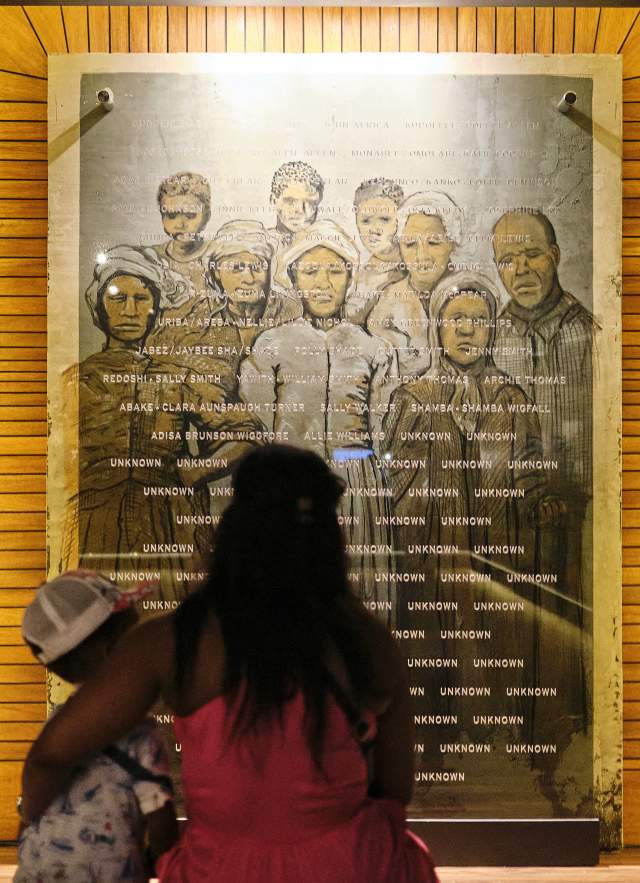 A Black woman with long hair holds her child while they look at a photo collage of formerly enslaved residents of Africatown, Alabama