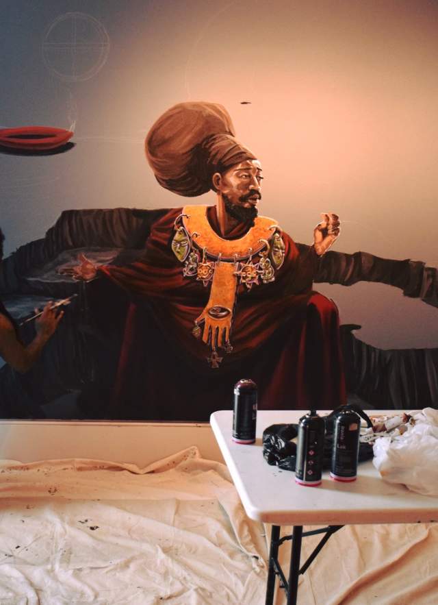 An artist kneels as he paints an African woman on a wall with artist supplies on a table