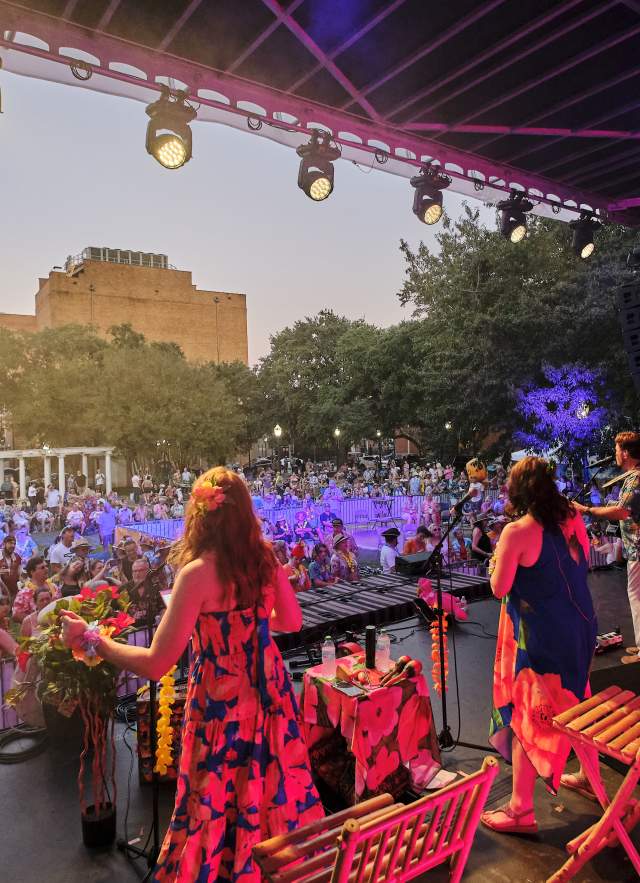 Group of singers and musicians dressed in tropical, summer attire performing in front of a crowd on stage