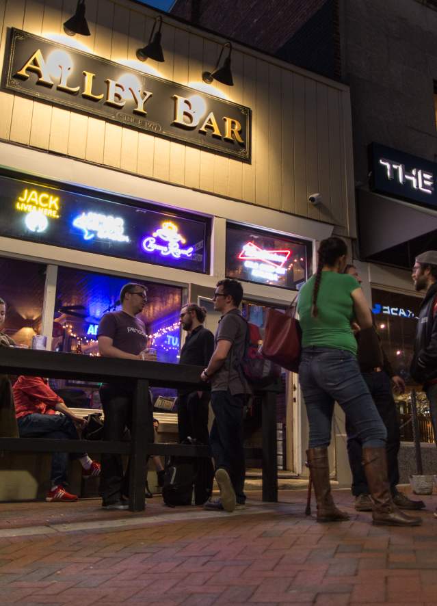 Alley Bar in Bloomington, Indiana at night, featuring a lively sidewalk scene with people gathered outside a bar with glowing signage, neon lights in the windows, and neighboring venue “The Tap Beer Company” visible along a brick-lined street.